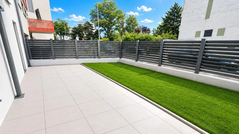 Große geflieste Terrasse mit Rasenfläche und modernem Zaun unter blauem Himmel.