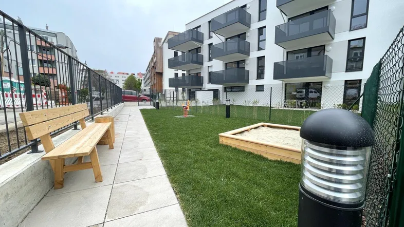 Gemeinschaftlicher Gartenbereich mit Rasenfläche, Sandkasten und Sitzbank vor einem modernen Wohngebäude.