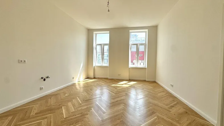 Helles, leeres Zimmer mit Fischgrätparkettboden und zwei großen Fenstern, bereit zur individuellen Gestaltung.