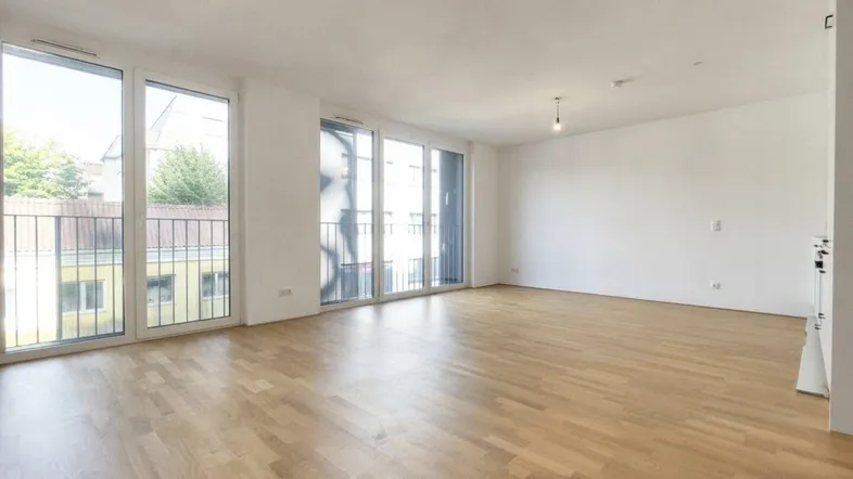 Helles, leeres Wohnzimmer mit großen Fenstern, Holzboden und Zugang zu einem französischen Balkon.