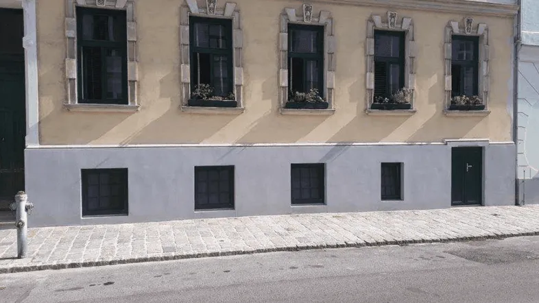 Klassische Fassade eines Altbaus mit verzierten Fenstern und einem gepflasterten Gehweg in der Sonne.