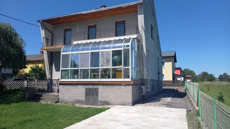 Hausfassade mit angebautem Wintergarten und Balkon im Obergeschoss, umgeben von Grünflächen.