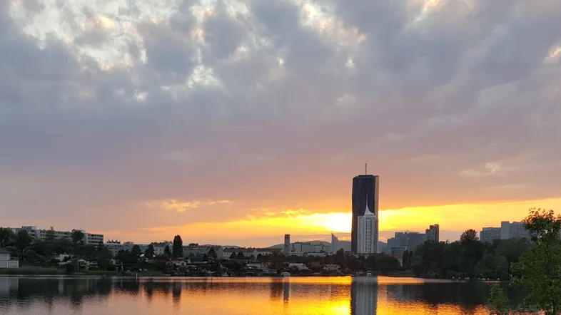 Sonnenuntergang über einem See mit Stadtpanorama und markantem Hochhaus, das sich im Wasser spiegelt.