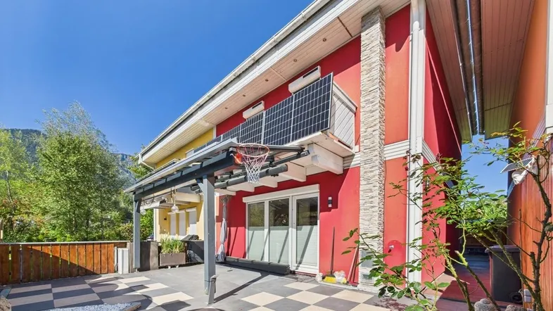 Rote Hausfassade mit gepflastertem Gartenbereich, Basketballkorb und Solarpaneelen auf dem Balkon.