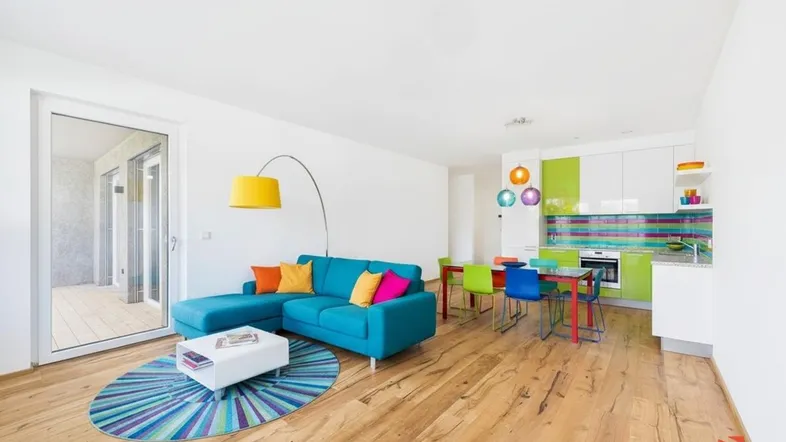 Helles, offenes Wohnzimmer mit bunter Küche, Holzboden und Zugang zum Balkon.