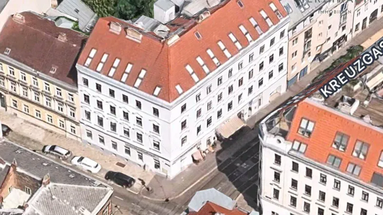 Luftaufnahme eines Eckgebäudes mit rotem Dach und vielen Dachfenstern in städtischer Umgebung.