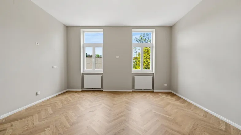 Helles, leeres Zimmer mit Fischgrätparkett und zwei großen Fenstern, ideal als Wohnzimmer nutzbar.