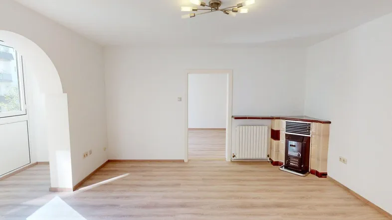 Helles Wohnzimmer mit Laminatboden, einem Kachelofen und Zugang zu einem weiteren Raum.