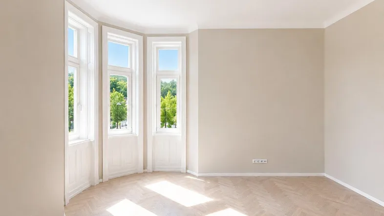 Helles, leeres Zimmer mit Fischgrätparkett und großen Fenstern, die viel Tageslicht hereinlassen.