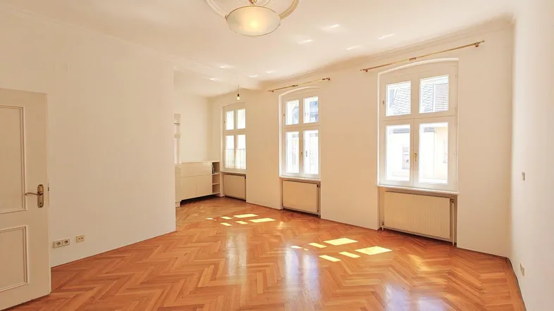 Helles Zimmer mit Fischgrätparkett, hohen Decken und großen Fenstern, ideal als Wohnbereich.