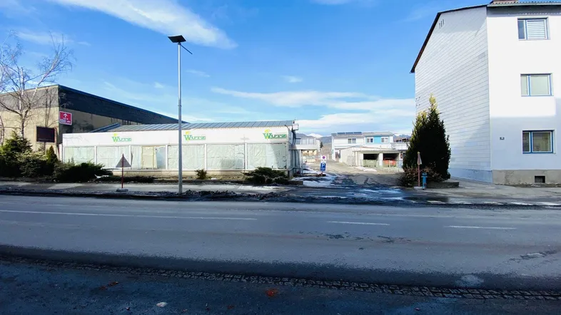 Blick von der Straße auf das Gewerbegebäude mit Parkplatz und umliegenden Wohngebäuden unter blauem Himmel.