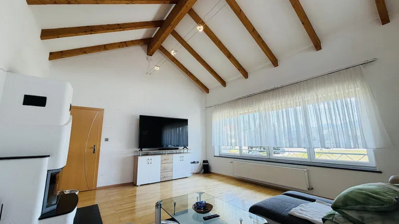 Helles Wohnzimmer mit Holzbalkendecke, Kaminofen und großen Fenstern mit Blick ins Grüne.