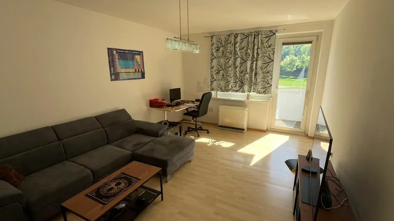 Helles Wohnzimmer mit gemütlichem Sofa, Arbeitsbereich und Zugang zum Balkon, ausgestattet mit hellem Laminatboden.