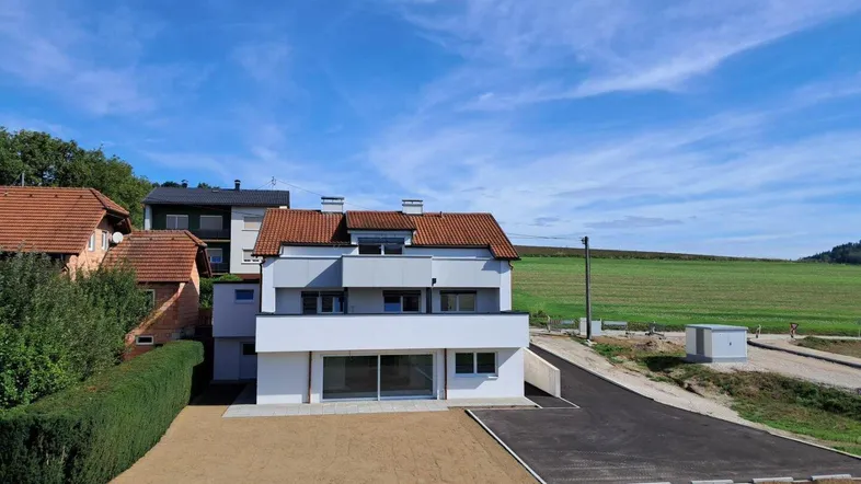 Modernes Wohnhaus mit weißer Fassade und rotem Ziegeldach, umgeben von grüner Landschaft und Feldern.