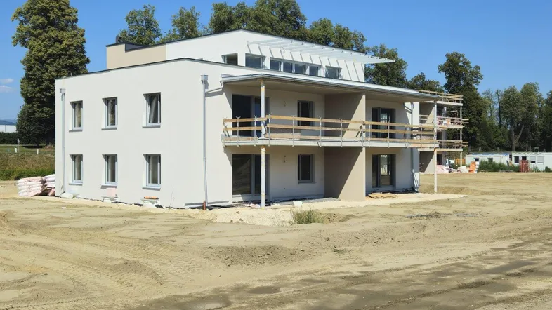 Modernes Mehrfamilienhaus im Bau mit Balkonen und umliegendem Baugrundstück unter blauem Himmel.