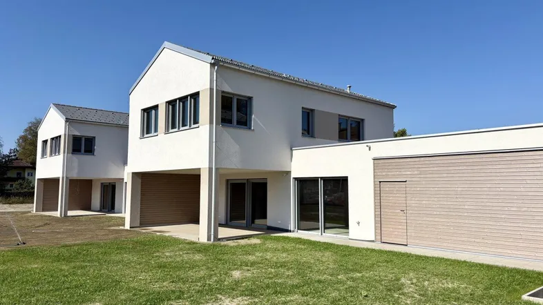 Zwei moderne Neubauhäuser mit weißer Fassade, großen Fenstern und einem grünen Rasen im Vordergrund.