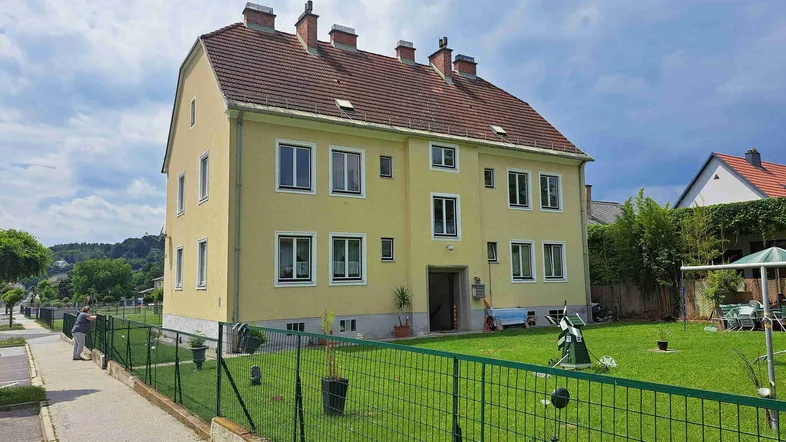 Gelbes Mehrfamilienhaus mit großem Gartenbereich und grüner Umzäunung unter blauem Himmel.