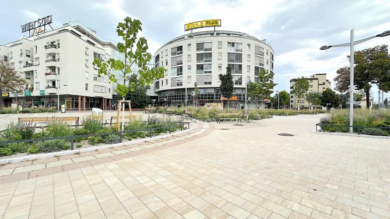 Moderner Stadtplatz mit gepflasterten Wegen und Gebäuden im Hintergrund unter bewölktem Himmel.