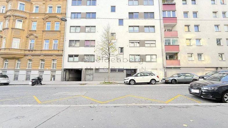 Mehrstöckiges Wohngebäude mit moderner Fassade und Parkplätzen an der Straße in urbaner Umgebung.
