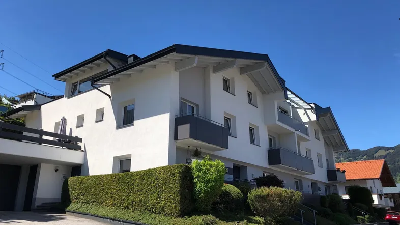 Modernes Mehrfamilienhaus mit weißer Fassade, Balkonen und gepflegter Hecke unter blauem Himmel.