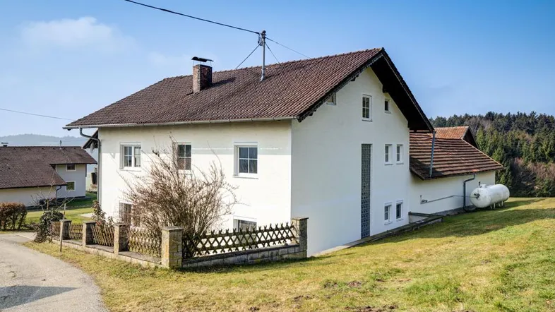 Freistehendes Einfamilienhaus mit weißer Fassade und braunem Ziegeldach, umgeben von grünen Hügeln und ländlicher Natur.