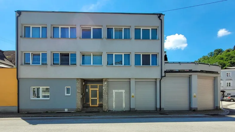 Gebäudefassade mit mehreren Fenstern, Haupteingang und zwei Garagentoren unter blauem Himmel.