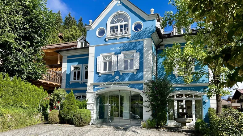 Historische Villa mit auffälliger blauer Fassade und weißen Fensterläden, umgeben von Grün.