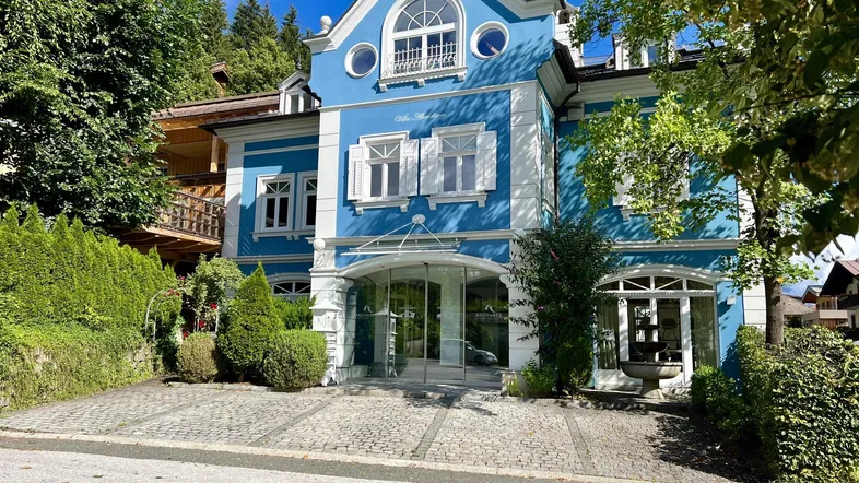 Prächtige blaue Villa mit weißer Fassadenverzierung und gepflastertem Vorplatz unter blauem Himmel.