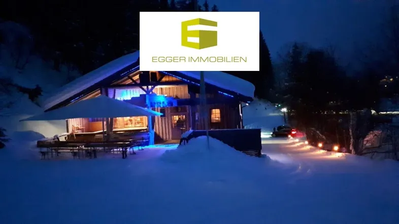 Gemütliches Holzgebäude im Schnee bei Nacht, beleuchtet mit blauen und warmen Lichtern, umgeben von verschneiter Landschaft.
