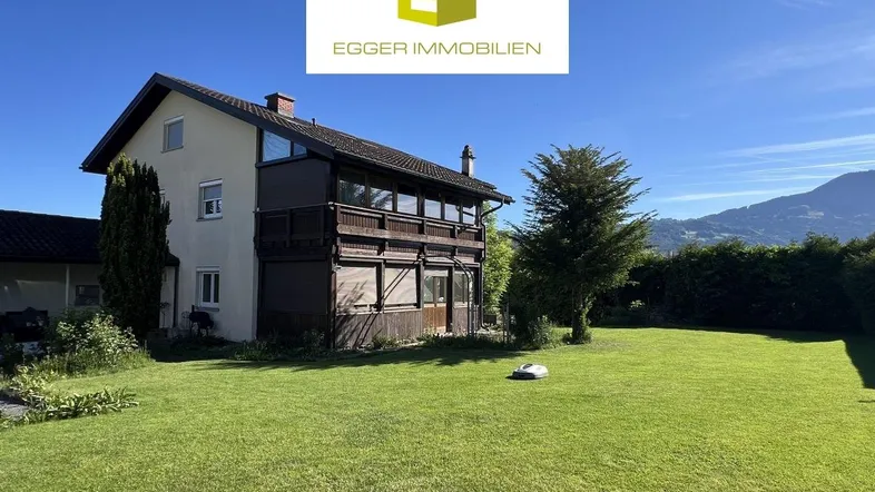 Großzügiger Gartenbereich mit Einfamilienhaus und Bergblick unter blauem Himmel.