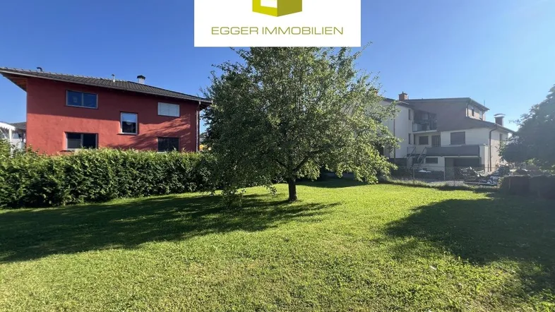 Großer, gepflegter Gartenbereich mit Rasenfläche und einem Baum, umgeben von Hecken und Wohnhäusern.