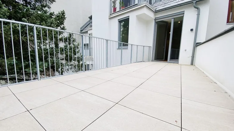 Große, geflieste Terrasse mit Metallgeländer und Zugang zu einem Gebäude mit Balkon.