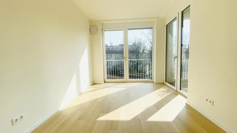 Helles Zimmer mit Parkettboden und Zugang zu einem Balkon, durchflutet von Sonnenlicht.