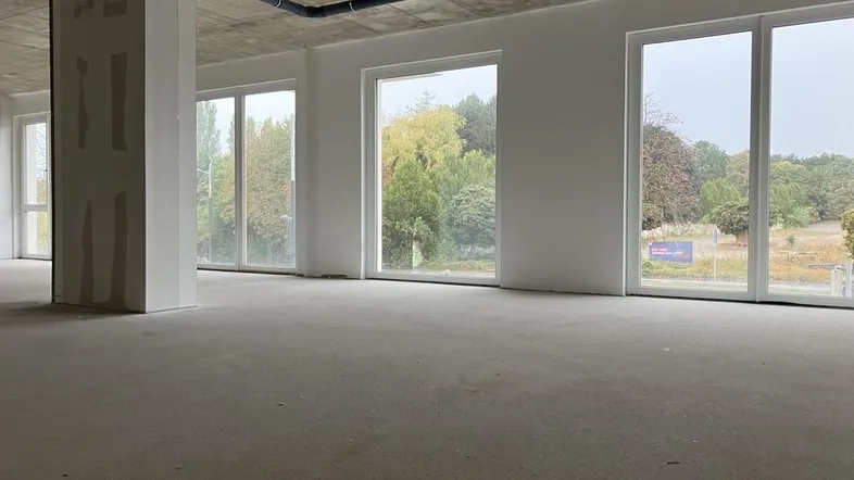 Großer, unfertiger Innenraum mit Betonboden und großen Fenstern, die viel natürliches Licht hereinlassen.
