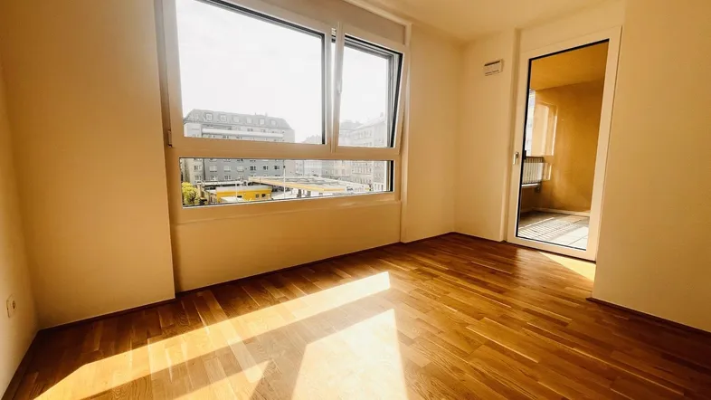 Helles Zimmer mit Parkettboden und großem Fenster, das viel Tageslicht hereinlässt und einen Stadtblick bietet.