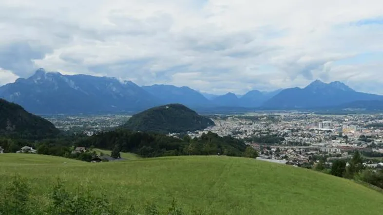 Weitläufiger Blick über eine grüne Landschaft mit einer Stadt im Tal und majestätischen Bergen im Hintergrund.