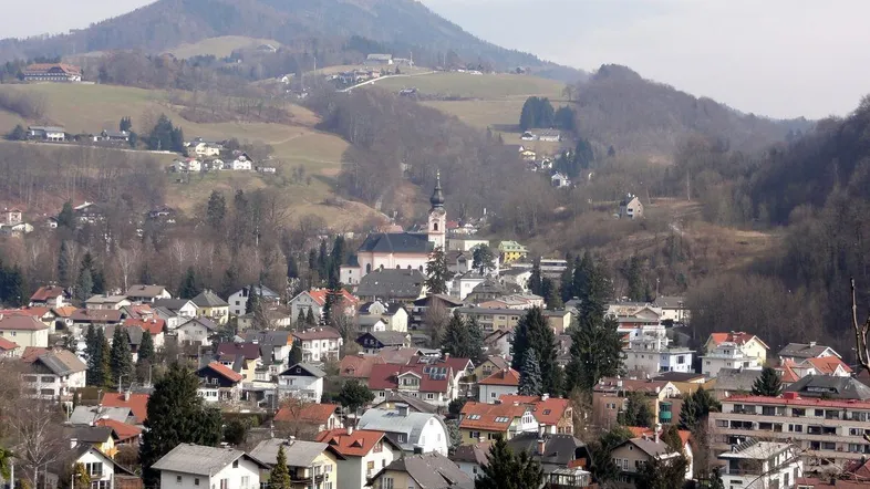 Panoramablick auf ein malerisches Dorf eingebettet in eine hügelige Landschaft mit bewaldeten Bergen im Hintergrund.
