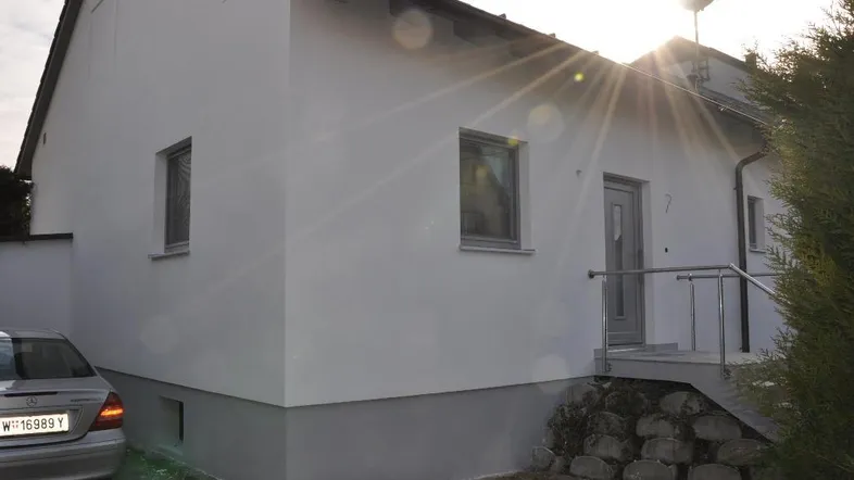 Modernes Einfamilienhaus mit weißer Fassade und grauem Sockel, sonniger Eingangsbereich mit Treppe.