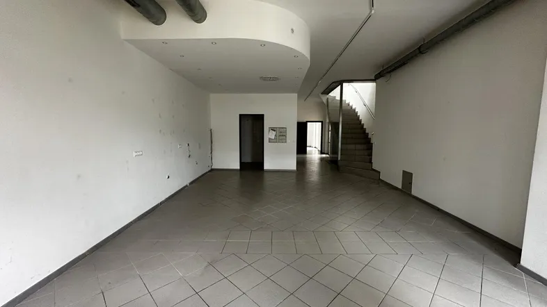 Geräumiger, leerer Gewerberaum mit Fliesenboden und sichtbaren Lüftungsrohren an der Decke.