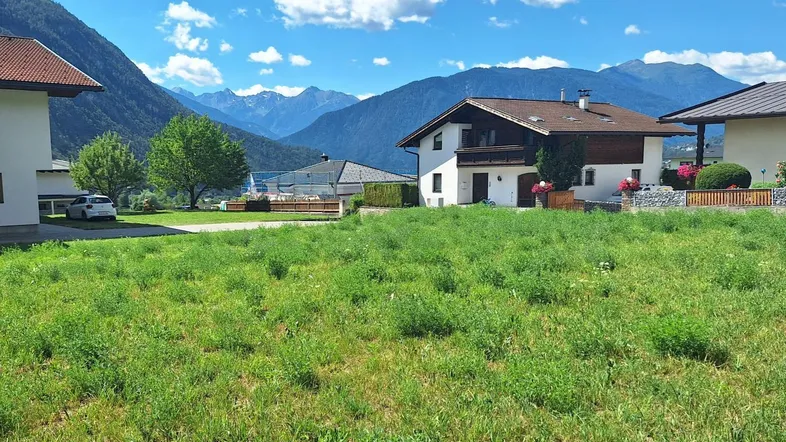 Grüne Wiese mit Blick auf Wohnhäuser und die umliegende Berglandschaft unter blauem Himmel.