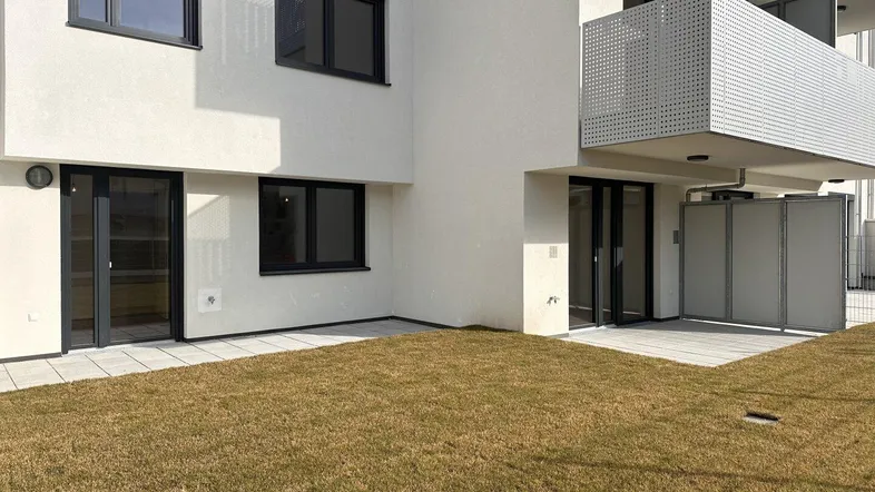 Gartenbereich mit Rasen und gepflasterter Terrasse, angrenzend an ein modernes Wohngebäude.