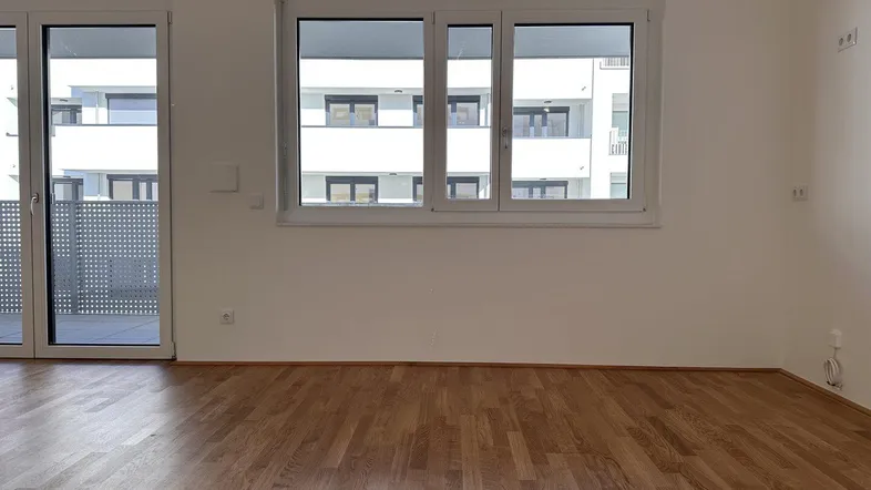 Helles Zimmer mit Parkettboden und Zugang zu einem Balkon, Blick auf andere Gebäude.