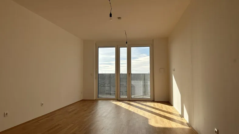 Geräumiges Wohnzimmer mit Holzboden und Zugang zu einem Balkon, hell durch Tageslicht.