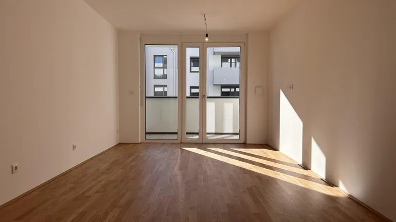 Helles, leeres Zimmer mit Holzboden und Zugang zu einem sonnigen Außenbereich.