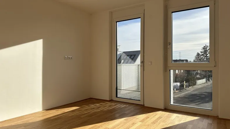 Helles, leeres Zimmer mit Parkettboden und Zugang zu einem Balkon, durchflutet von Sonnenlicht.