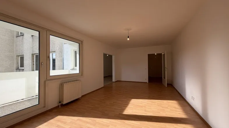 Helles, leeres Zimmer mit Laminatboden und großen Fenstern, das viel Tageslicht hereinlässt.