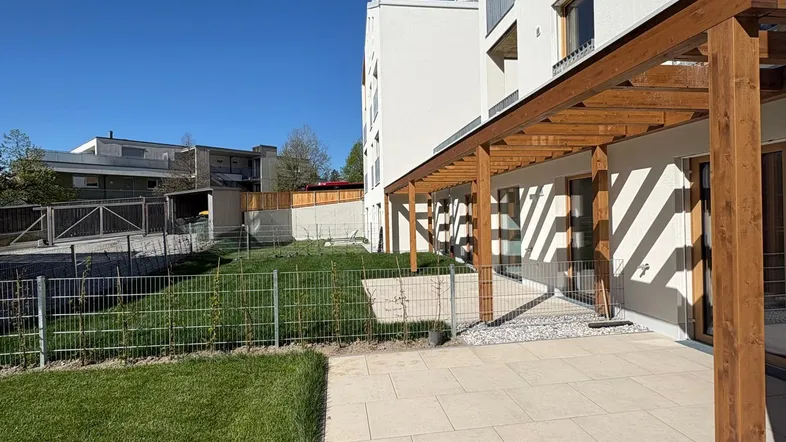 Moderne Terrasse mit Holzpergola und angrenzender Rasenfläche vor einem Neubaugebäude.