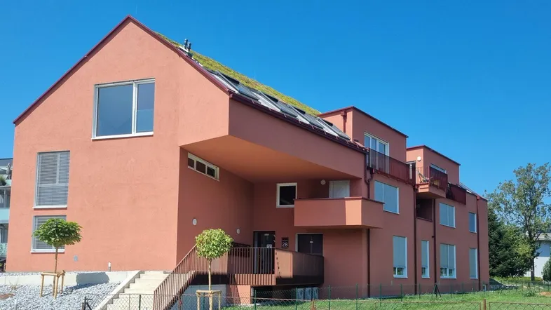 Modernes Wohngebäude mit roter Fassade, Gründach und Balkonen unter blauem Himmel.