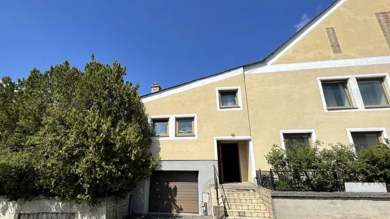 Gelbe Hausfassade mit Garagentor, Eingangsbereich und grünen Büschen unter blauem Himmel.