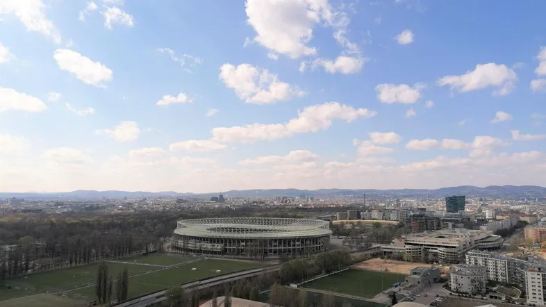 Weitläufiger Blick über die Stadt mit dem Stadion und umliegenden Grünflächen unter blauem Himmel.